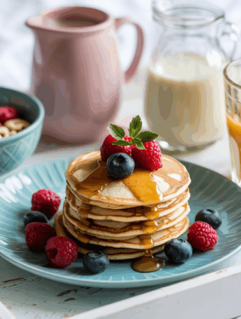 Breakfast in bed featuring a stack of pancakes topped with fresh raspberries and blueberries, drizzled with syrup on a blue plate. Accompanied by a bowl of cereal, a glass of orange juice, a jug of milk, and a pink flower on a white tray.