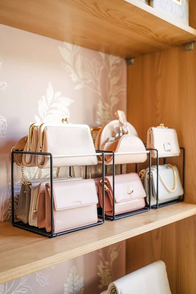A beautifully organized collection of small handbags and purses displayed on a wooden shelf. The bags are arranged in black metal organizer cubes that keep them neatly separated and visible. The collection features several elegant pastel-colored purses in light pink and cream tones, many with gold chain straps and clasps. The wooden shelving unit has a decorative floral patterned backing, creating an elegant boutique-like display. This storage solution effectively showcases the purse collection while keeping everything accessible and preventing the bags from losing their shape.