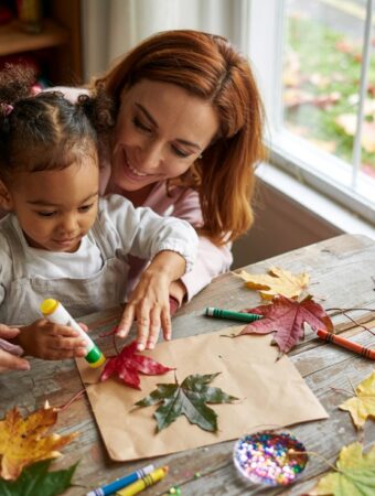 A woman and young child doing leaf crafts together at a wooden table by a window with colorful fall leaves, demonstrating simple fall activities for kids at home.
