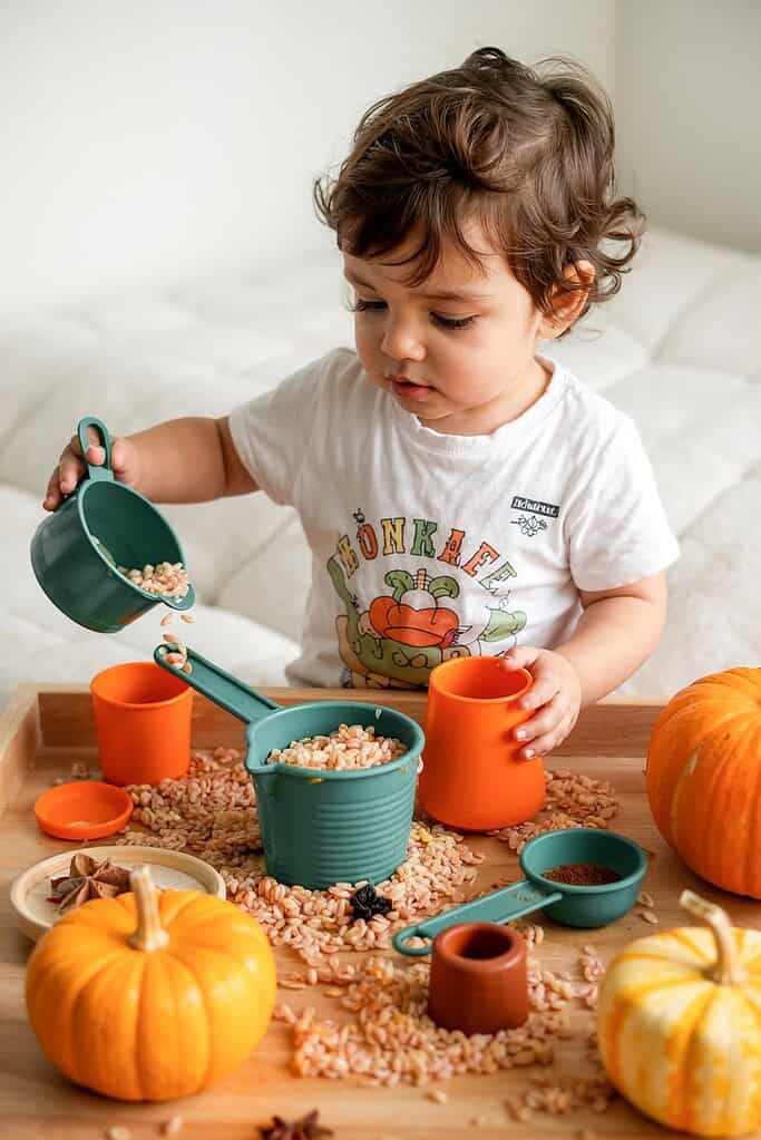 Child in pumpkin shirt playing with fall-themed sensory bin, scooping rice with measuring cups surrounded by decorative pumpkins on wooden tray.
