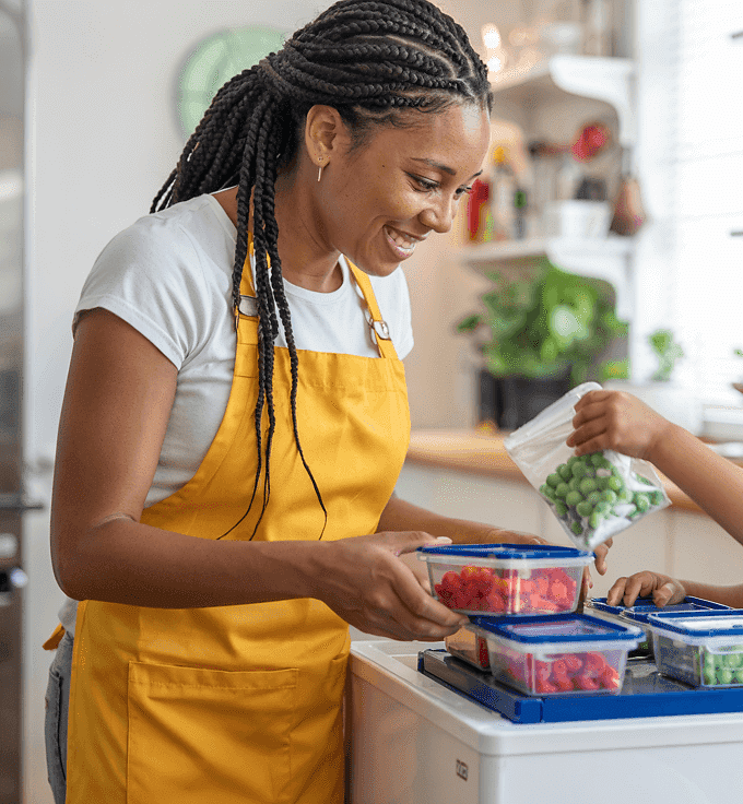A woman and a child are engaged in meal preparation in a kitchen. The woman is wearing a yellow apron and organizing food in containers, while the child is helping by adding green vegetables to one of the containers. The kitchen is well-stocked with various ingredients and utensils visible in the background.