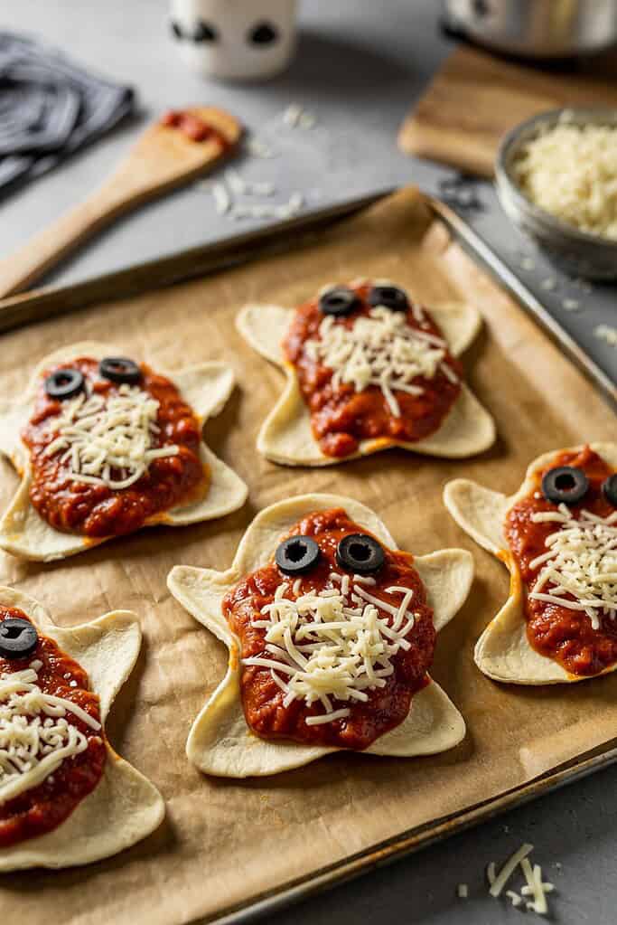 Tray of baked mini ghost-shaped pizzas with cheese faces and olives for eyes, surrounded by small pumpkins.