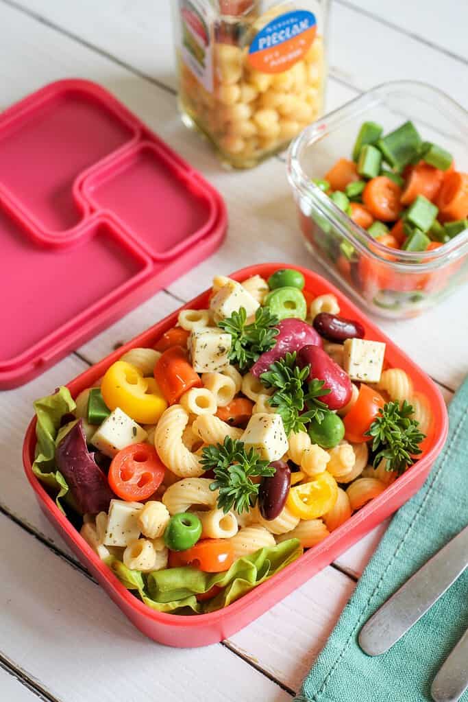 Colorful pasta salad in a pink lunchbox with fresh vegetables and herbs.