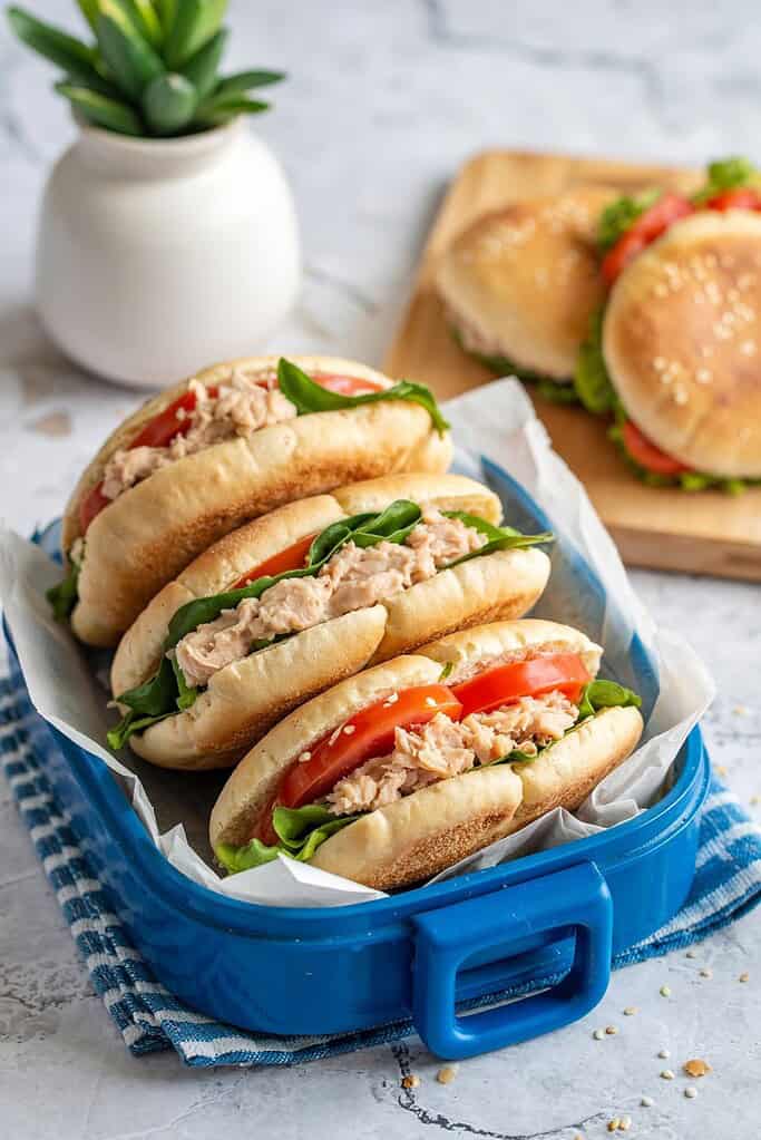 Fresh tuna salad sandwiches in a blue lunchbox with tomatoes and greens.