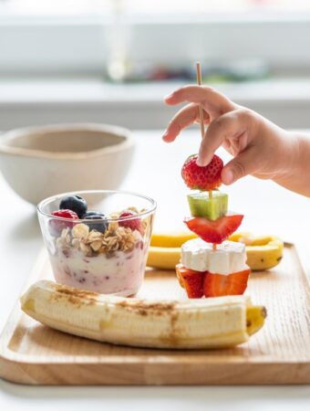 no cook breakfast toddler meal showing a hand holding a skewer of fruit and yogurt